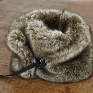May include: A brown faux fur collar with a black strap and a silver buckle. The collar is folded in half and the strap is looped through the buckle.