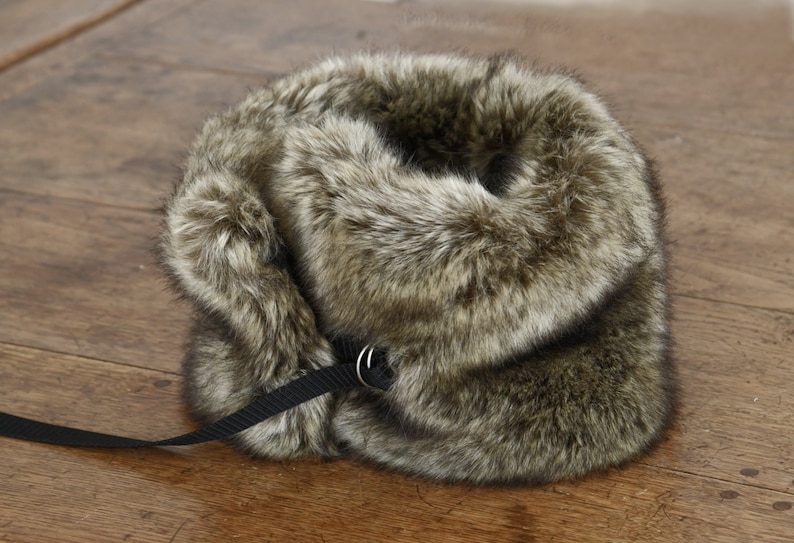 May include: A brown faux fur collar with a black strap and a silver buckle. The collar is folded in half and the strap is looped through the buckle.