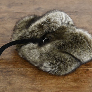 May include: A brown and gray fur hat with a black strap and a silver buckle. The hat is lying on a wooden surface.