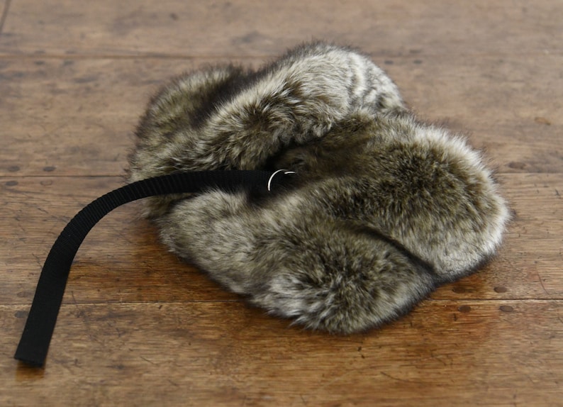 May include: A brown and gray fur hat with a black strap and a silver buckle. The hat is lying on a wooden surface.
