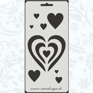 Pode incluir: Um estêncil com um desenho de vários corações, incluindo um coração grande com círculos concêntricos e corações menores espalhados ao redor. O estêncil é feito de um material branco e é adequado para projetos de artesanato.