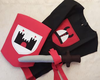 Puede incluir: Un disfraz de caballero rojo y negro para niños que incluye una capa, un escudo y una espada. El escudo tiene un diseño de castillo blanco y la capa tiene un parche rojo con un diseño de casco negro.