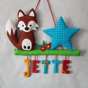 Peut inclure: Décoration murale en feutre avec un renard marron et une étoile bleue à pois blancs. Le prénom "JETTE" est écrit en lettres de feutre colorées, suspendues à une branche verte avec une ficelle rouge.