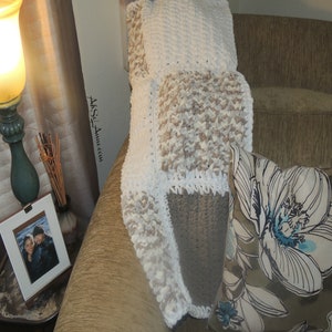 May include: A white and beige crocheted throw blanket with a square pattern. The blanket is draped over a brown couch with a floral patterned pillow.