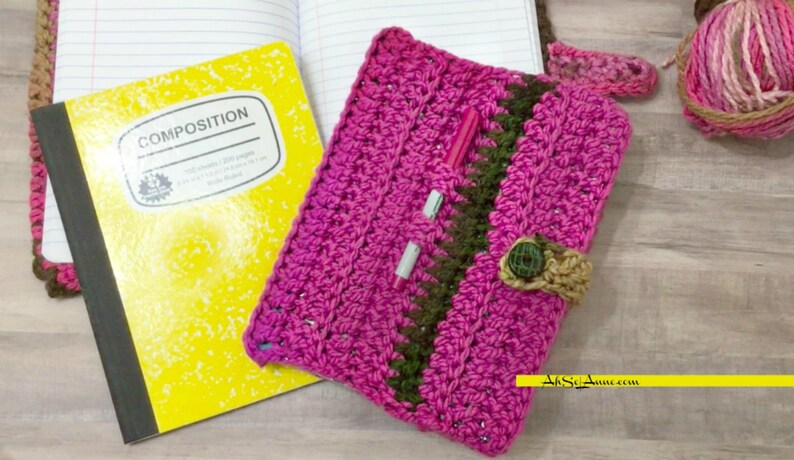 May include: A pink crocheted notebook cover with a green and brown stripe, a button closure, and a pen tucked inside. The cover is lying on top of a yellow composition notebook.