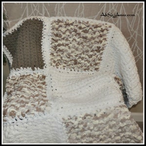 May include: A crocheted throw blanket with a patchwork design in shades of white, brown, and beige. The blanket is made of chunky yarn and has a soft, fluffy texture.