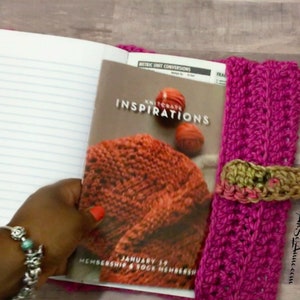 May include: A pink crocheted notebook cover with a brown crocheted closure. The cover is holding a magazine titled "Knitcrate Inspirations" with a photo of a knitted item on the front. The magazine has the text "January 19 Membership & Sock Membership" on the bottom.