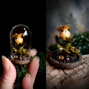 Könnte beinhalten: Eine Miniatur-Pilz-Kreatur-Skulptur unter einer Glaskuppel. Die Skulptur zeigt eine kleine, orange-braune pilzartige Figur mit einem süßen Ausdruck, eingebettet in grünes und goldenes Laub. Der Sockel ist ein dunkelbrauner Holzring.