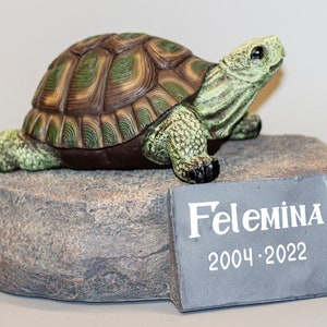 Puede incluir: Una pequeña figura de tortuga verde y marrón sobre una base de roca gris. Una placa de pizarra con la inscripción "Felemina 2004-2022" está unida a la base.