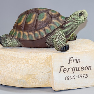 May include: A green and brown turtle figurine on a beige rock base with a plaque that reads "Erin Ferguson 1900-1973".