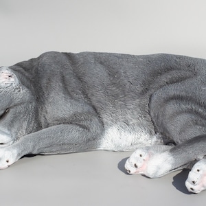 Peut inclure: Une figurine de chat gris et blanc endormi. Le chat est couché sur le côté, la tête reposant sur ses pattes. La figurine est faite d'un matériau réaliste et a une peinture détaillée.