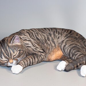 May include: A ceramic sculpture of a tabby cat sleeping on its side. The cat is grey and black with white paws and a white chest.