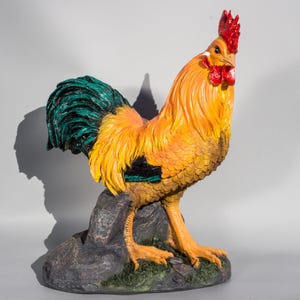 Könnte beinhalten: Eine gelbe und grüne Hahnfigur mit rotem Kamm und Kehllappen. Der Hahn steht auf einem grauen Steinsockel.