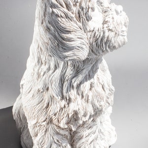May include: White ceramic sculpture of a dog sitting with its head tilted to the side. The dog has a fluffy, textured coat and a sweet expression.
