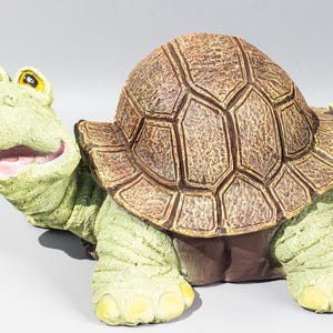 Pode incluir: Uma figura de tartaruga caprichosa com corpo verde claro e carapaça marrom. A tartaruga tem um rosto sorridente com olhos amarelos e boca rosa. Enfeite decorativo para jardim.