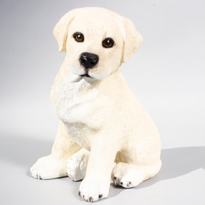 May include: A white Labrador Retriever puppy figurine sitting with its paws crossed. The figurine is made of a realistic-looking material and has a sweet expression on its face.