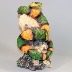 May include: A green and orange snake figurine coiled around a tree stump. The snake has a realistic texture and is painted with bright colors.