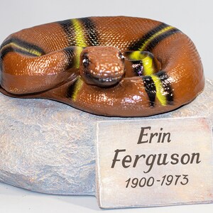 Könnte beinhalten: Eine braune und gelbe Schlangenfigur auf einem grauen Steinsockel mit einer Plakette mit der Aufschrift "Erin Ferguson 1900-1973".