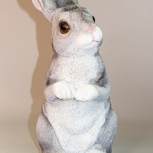May include: A gray and white rabbit figurine with brown eyes. The rabbit is sitting upright with its paws crossed in front of it.