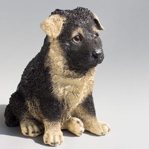 May include: A black and tan German Shepherd puppy figurine sitting with its head tilted to the side. The figurine is made of a realistic-looking material and has a detailed face.
