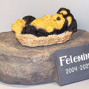 May include: A small, black and tan dog figurine is lying in a wicker basket on a gray rock. A black slate plaque with the name "Felemina" and the dates "2004-2022" is on the rock.