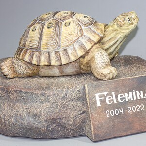 Puede incluir: Una pequeña figura de tortuga de cerámica marrón sobre una base de roca con una placa que dice "Felemina 2004-2022".