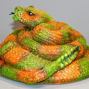 Könnte beinhalten: Eine gewickelte grüne und orangefarbene Klapperschlangenfigur. Die Schlange ist zusammengerollt, mit dem Kopf auf dem Körper liegend. Die Figur ist aus einem strukturierten Material gefertigt, das ihr ein realistisches Aussehen verleiht.