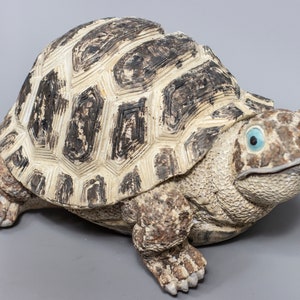 Puede incluir: Una figurita de tortuga de cerámica con una concha marrón y blanca y ojos azules. La tortuga está en posición de caminar con la cabeza girada hacia un lado.