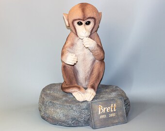 May include: A brown and white monkey figurine sitting on a gray rock base. The base has a small plaque with the name "Brett" and the dates "1993 - 2022" inscribed on it.