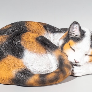 Puede incluir: Figura de gato de cerámica en posición de dormir enrollada. El gato tiene un patrón de calicó con pelaje negro, naranja y blanco. Los ojos del gato están cerrados y sus patas están metidas.