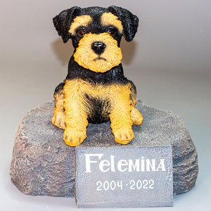 May include: A small black and tan dog figurine sitting on a gray rock base. A plaque on the base reads "Felemina 2004-2022".