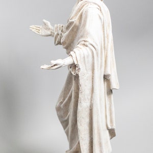 May include: White painted figurine of a person with long hair and a flowing robe, standing with arms outstretched.