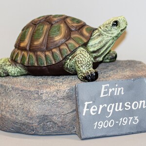 May include: A green and brown turtle figurine on a grey rock base with a slate plaque that reads "Erin Ferguson 1900-1973".
