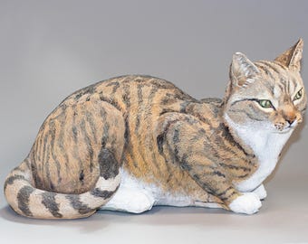May include: A realistic ceramic sculpture of a tabby cat lying down. The cat has brown, black, and white fur with green eyes. The sculpture is detailed with realistic fur and features.