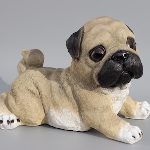 May include: A beige and white pug puppy figurine with black eyes and a black nose. The figurine is lying on its belly with its paws outstretched.
