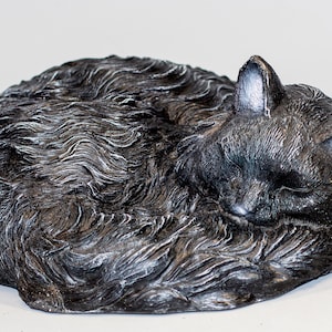May include: A sleeping cat figurine in a curled position. The cat is dark gray with silver highlights, and its fur has a textured, wavy appearance. The cat's eyes are closed, and its ears are perked up.