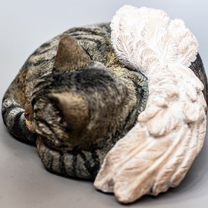 Op de afbeelding: Een grijs en bruin tabby kattenbeeldje met een witte gevederde engelenvleugel. De kat ligt opgerold in een slaaphouding.