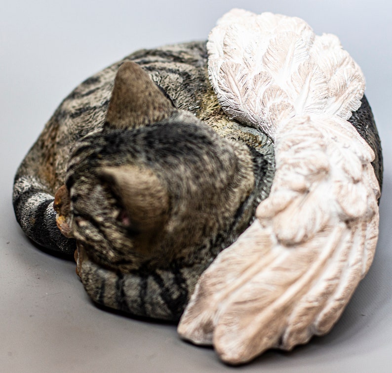 Op de afbeelding: Een grijs en bruin tabby kattenbeeldje met een witte gevederde engelenvleugel. De kat ligt opgerold in een slaaphouding.