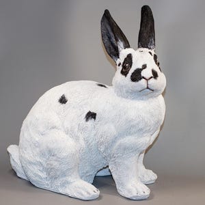 Può includere: Una statuetta di coniglio bianco con macchie nere e orecchie. Il coniglio è seduto con la testa girata di lato e guarda lo spettatore.