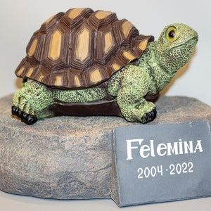 Puede incluir: Una pequeña figura de tortuga verde y marrón sobre una base de roca gris con una placa de pizarra que dice "Felemina 2004-2022".