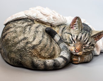 May include: A sleeping tabby cat figurine with white angel wings. The cat is curled up with its tail wrapped around its body. The figurine is made of a light brown and gray material.
