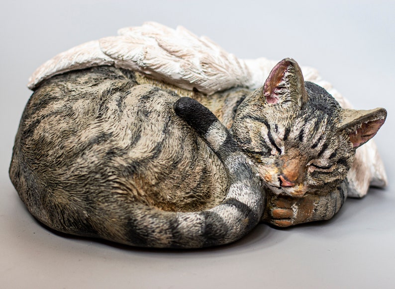 Op de afbeelding: Een slapende tabby kattenfiguur met witte engelenvleugels. De kat is opgerold met zijn staart om zijn lichaam gewikkeld. Het beeldje is gemaakt van lichtbruin en grijs materiaal.