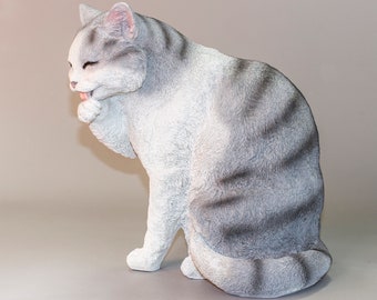 May include: A white and gray cat figurine with black stripes, sitting on its haunches and licking its paw.