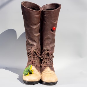 May include: A pair of brown ceramic boots with white tops. One boot has a green frog figurine on the toe and the other has a red ladybug figurine on the side.