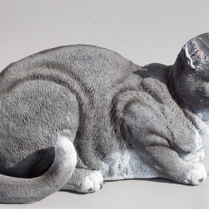 May include: A grey and white ceramic cat figurine with yellow eyes. The cat is lying down with its tail curled around its body.