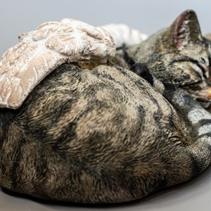 Op de afbeelding: Een slapende tabby kattenfiguur met een witte engelenvleugel op zijn rug. Het beeldje is gemaakt van geverfd hars en heeft een realistisch uiterlijk.