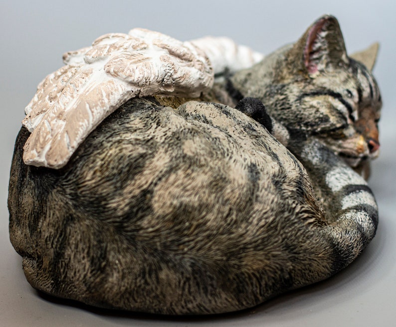 Op de afbeelding: Een slapende tabby kattenfiguur met een witte engelenvleugel op zijn rug. Het beeldje is gemaakt van geverfd hars en heeft een realistisch uiterlijk.