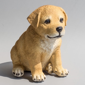 Puede incluir: Una pequeña figura realista de un cachorro sentado, de color marrón claro, con la cabeza inclinada hacia abajo. El cachorro tiene una mancha blanca en el pecho y ojos negros.