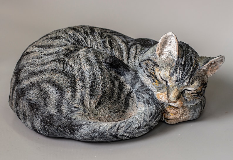 May include: A ceramic sculpture of a tabby cat curled up sleeping. The cat is gray and white with black stripes. The sculpture is realistic and detailed.