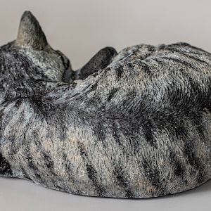 May include: A gray and white tabby cat sculpture curled up in a sleeping position. The sculpture is made of a textured material that resembles fur.
